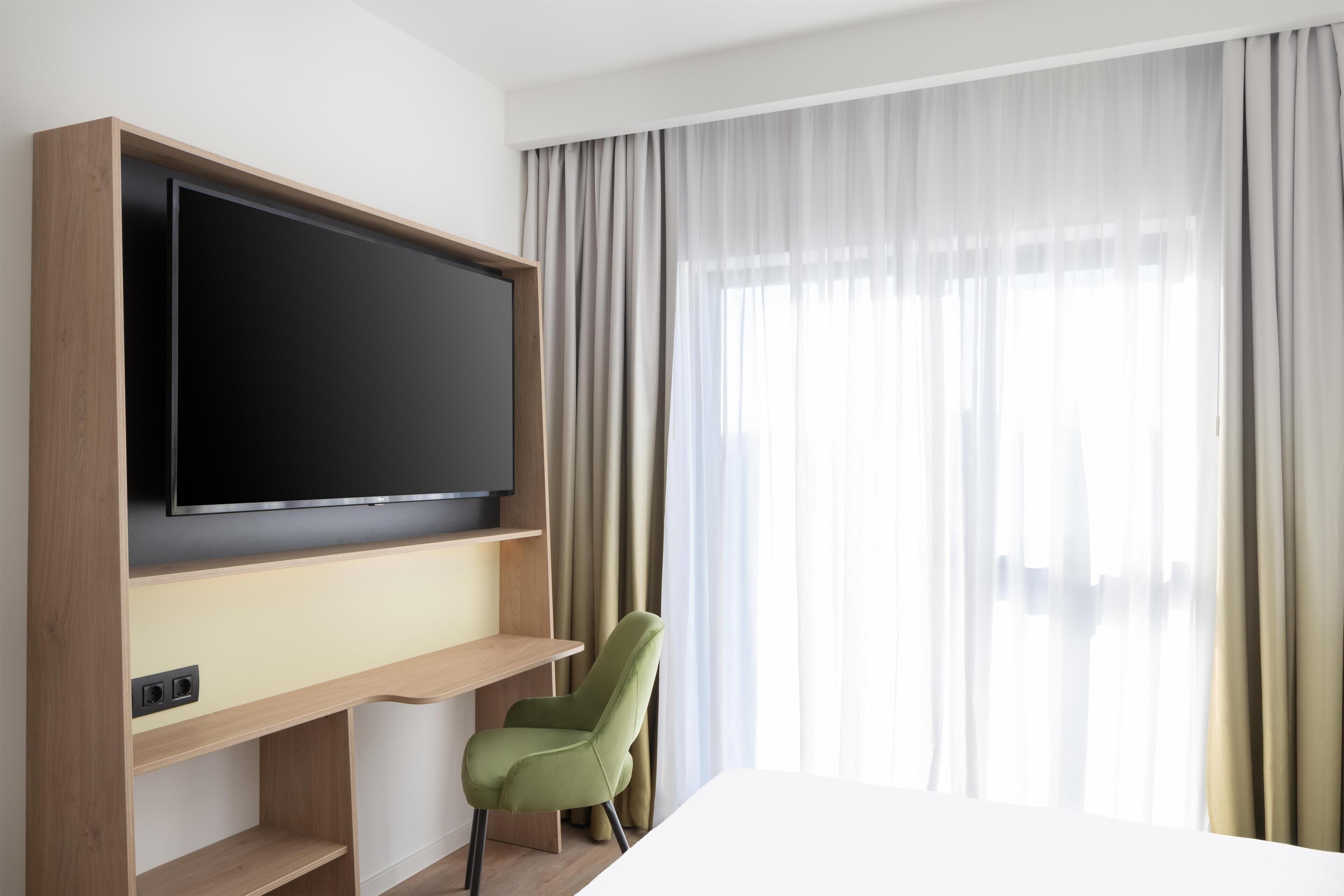 Habitación de hotel con un televisor de pantalla plana grande montado en un estante de madera, una silla verde y un ventanal con cortinas traslúcidas.