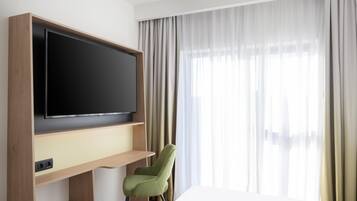 Habitación de hotel con un televisor de pantalla plana grande montado en un estante de madera, una silla verde y un ventanal con cortinas traslúcidas.