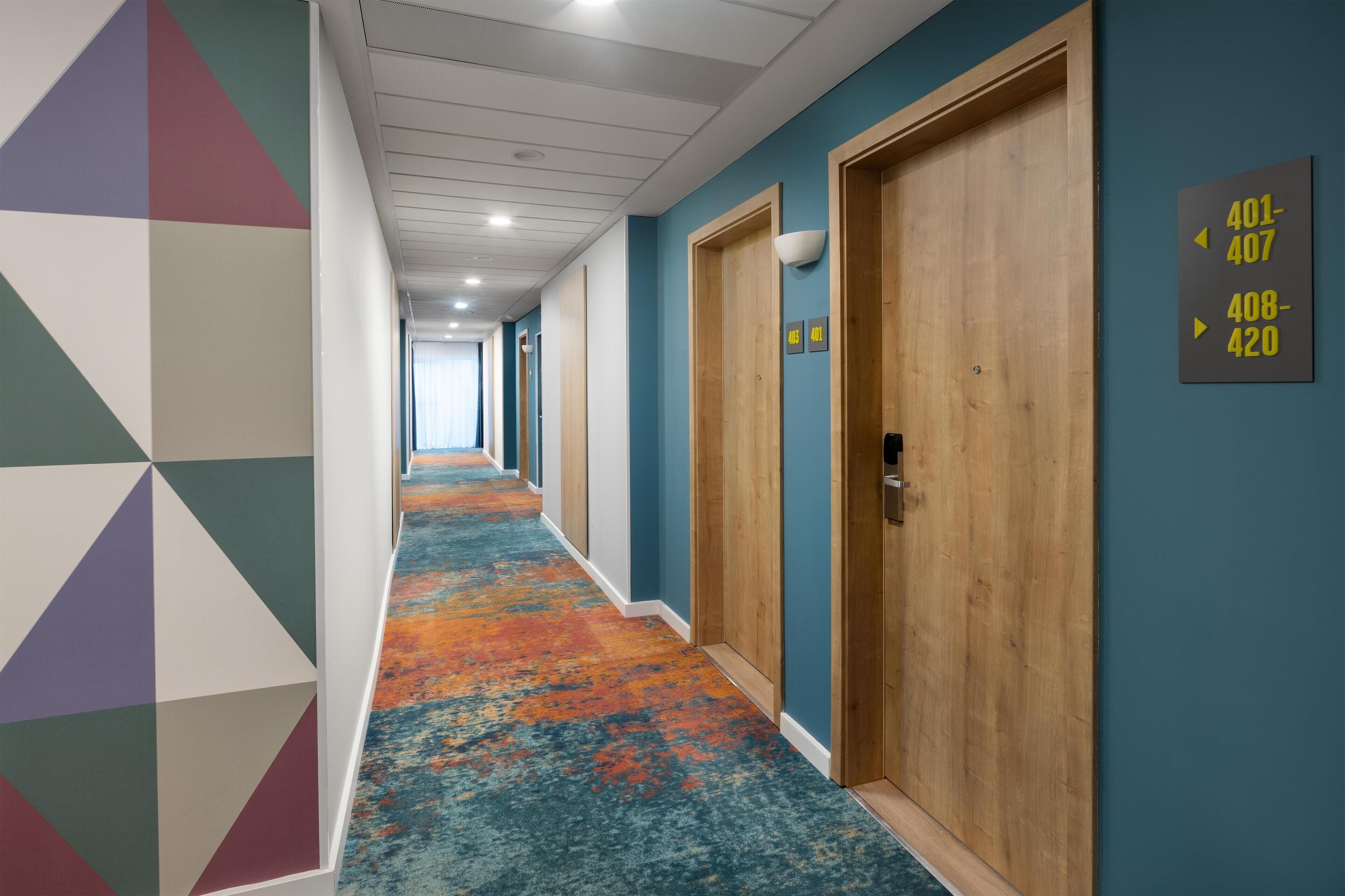 Un pasillo de hotel con arte mural geométrico y colorido, puertas de madera y piso alfombrado.