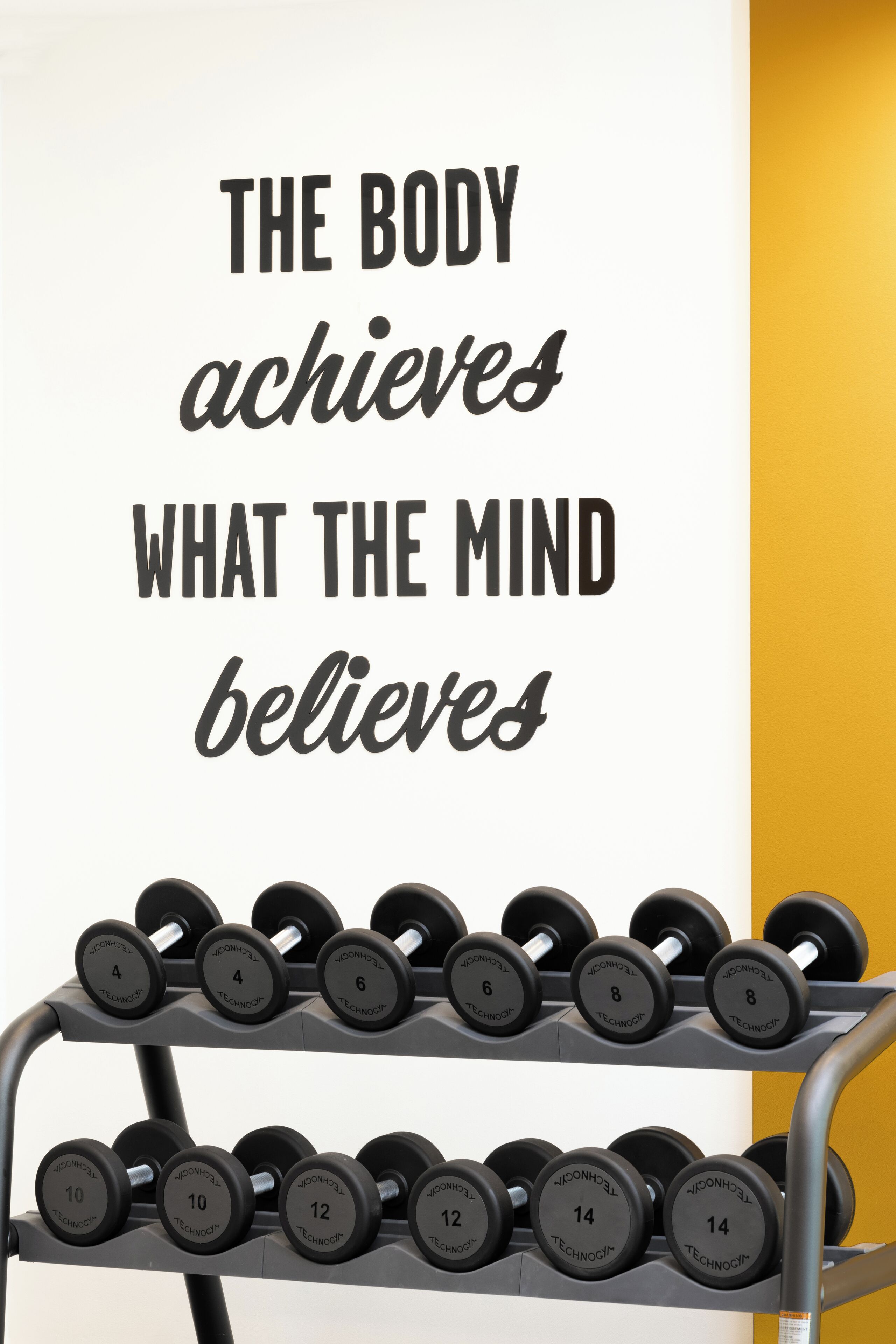 A motivational quote on a white wall with a black background. In front of the quote, there is a rack of black dumbbells with weights ranging from 4 to 14.