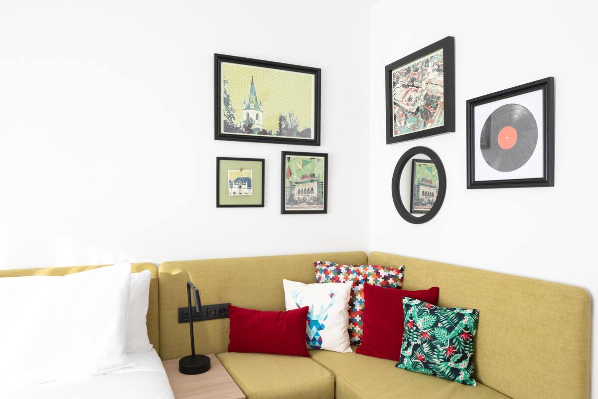A room with a yellow sofa, various framed artworks on the wall, and multiple pillows.