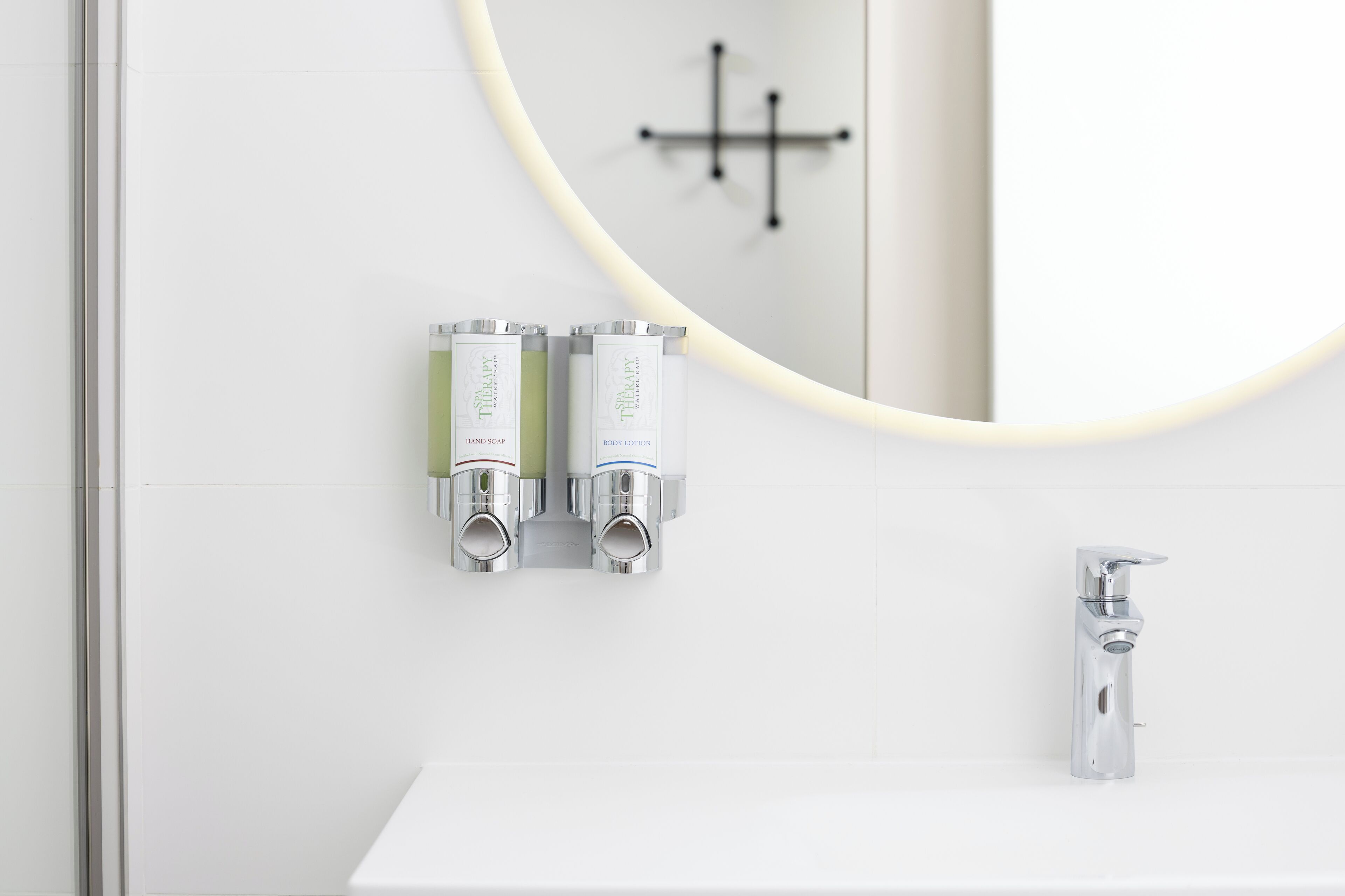 Un baño moderno con un lavamanos blanco, dos dispensadores de jabón líquido para manos, un dispensador de crema corporal y un grifo cromado.