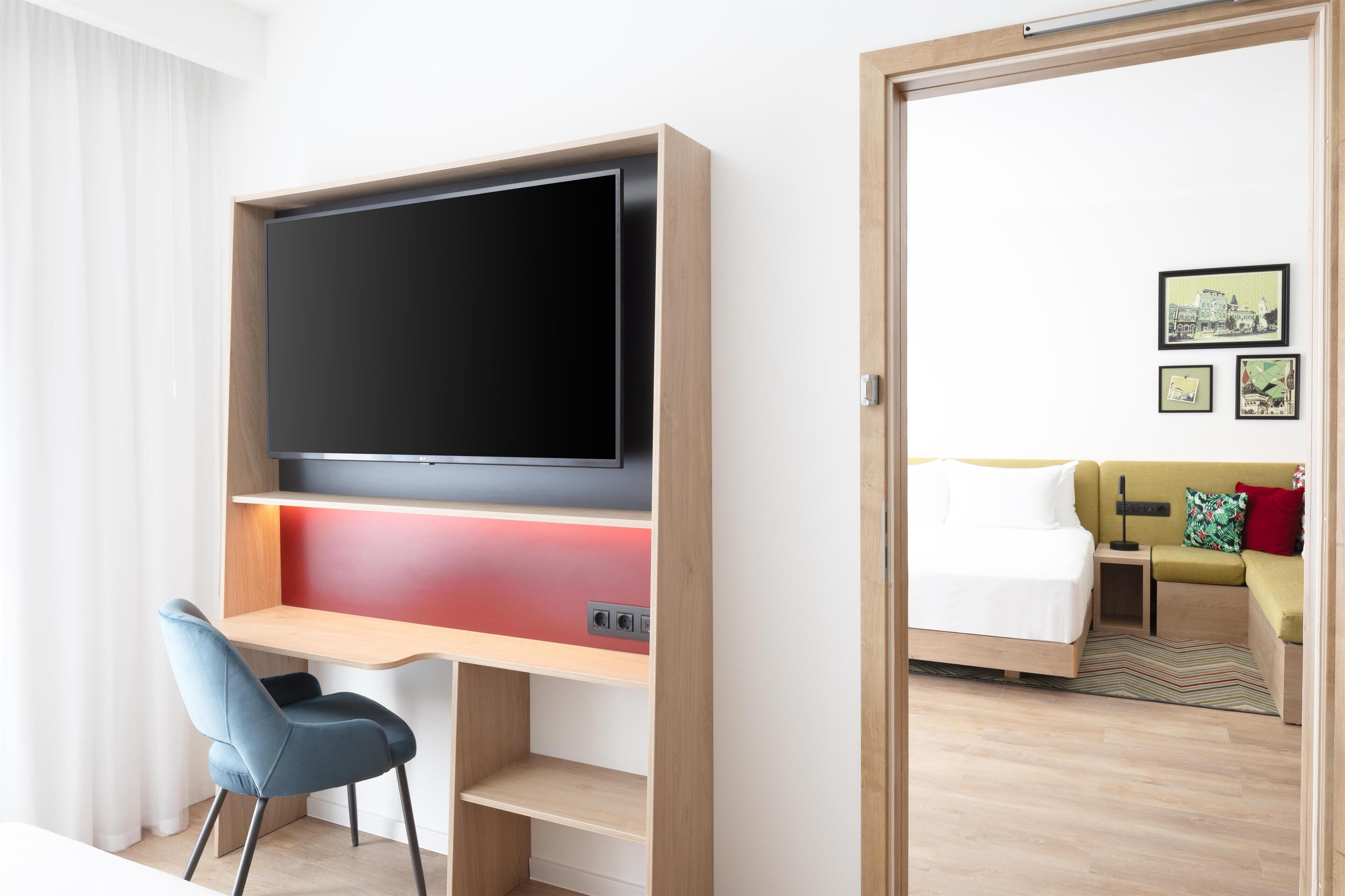 Habitación de hotel con televisión sobre un soporte de madera, un escritorio con silla, una cama y un sofá.