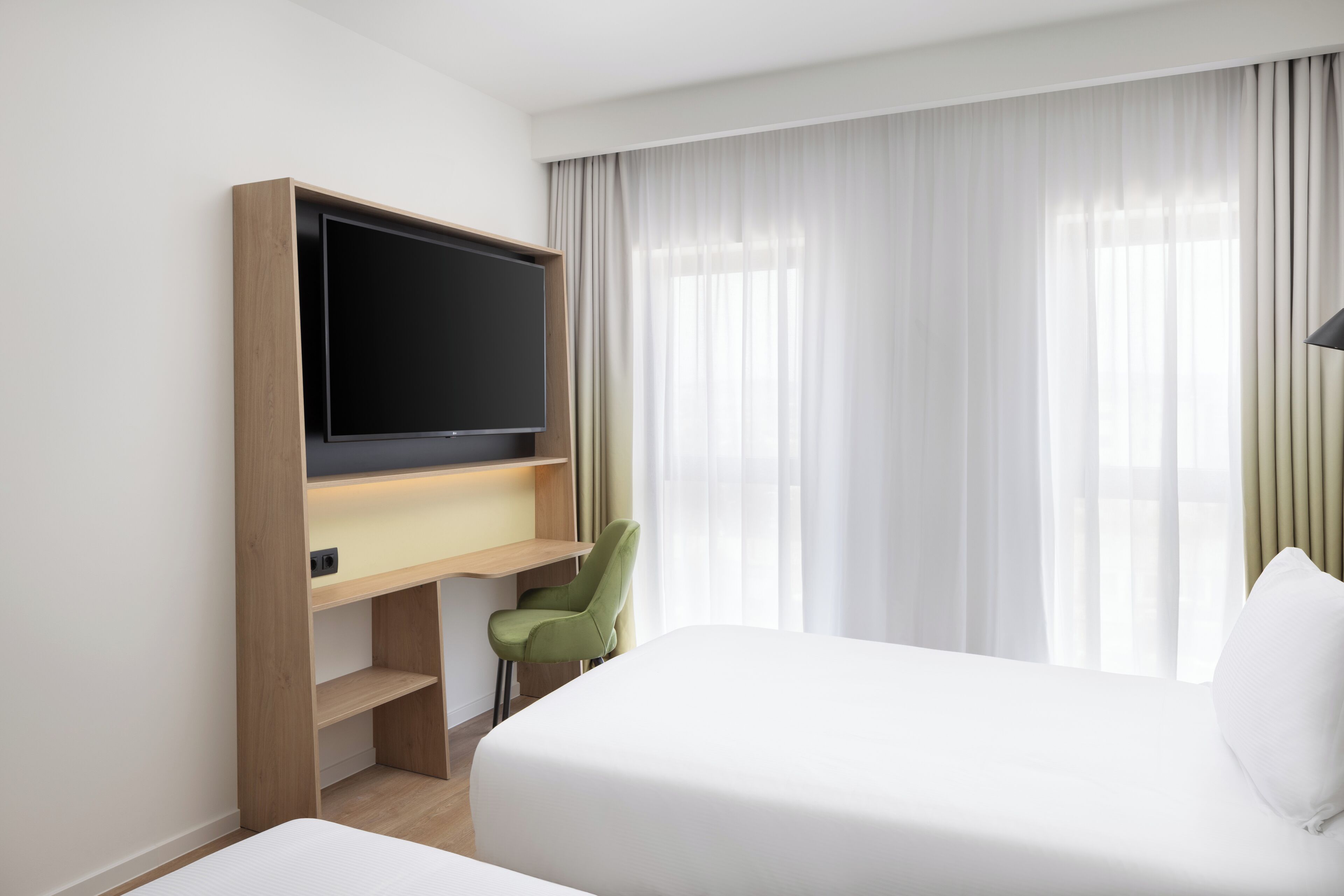 A hotel room with a bed, a desk with a flat-screen TV, a green chair, and a large window with sheer curtains.
