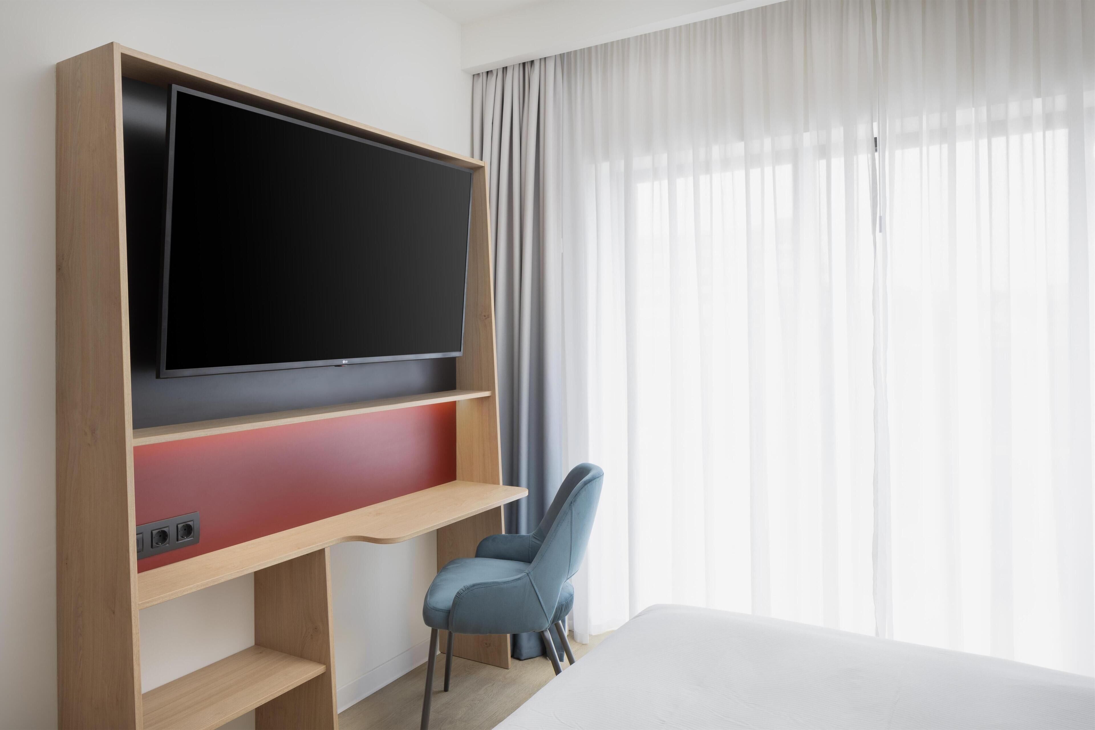 A hotel room with a large flat-screen TV mounted on a wooden wall, a blue chair, and a bed with white bedding.