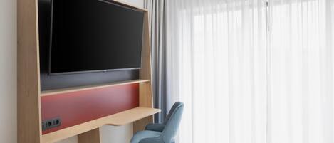 A hotel room with a large flat-screen TV mounted on a wooden wall, a blue chair, and a bed with white bedding.