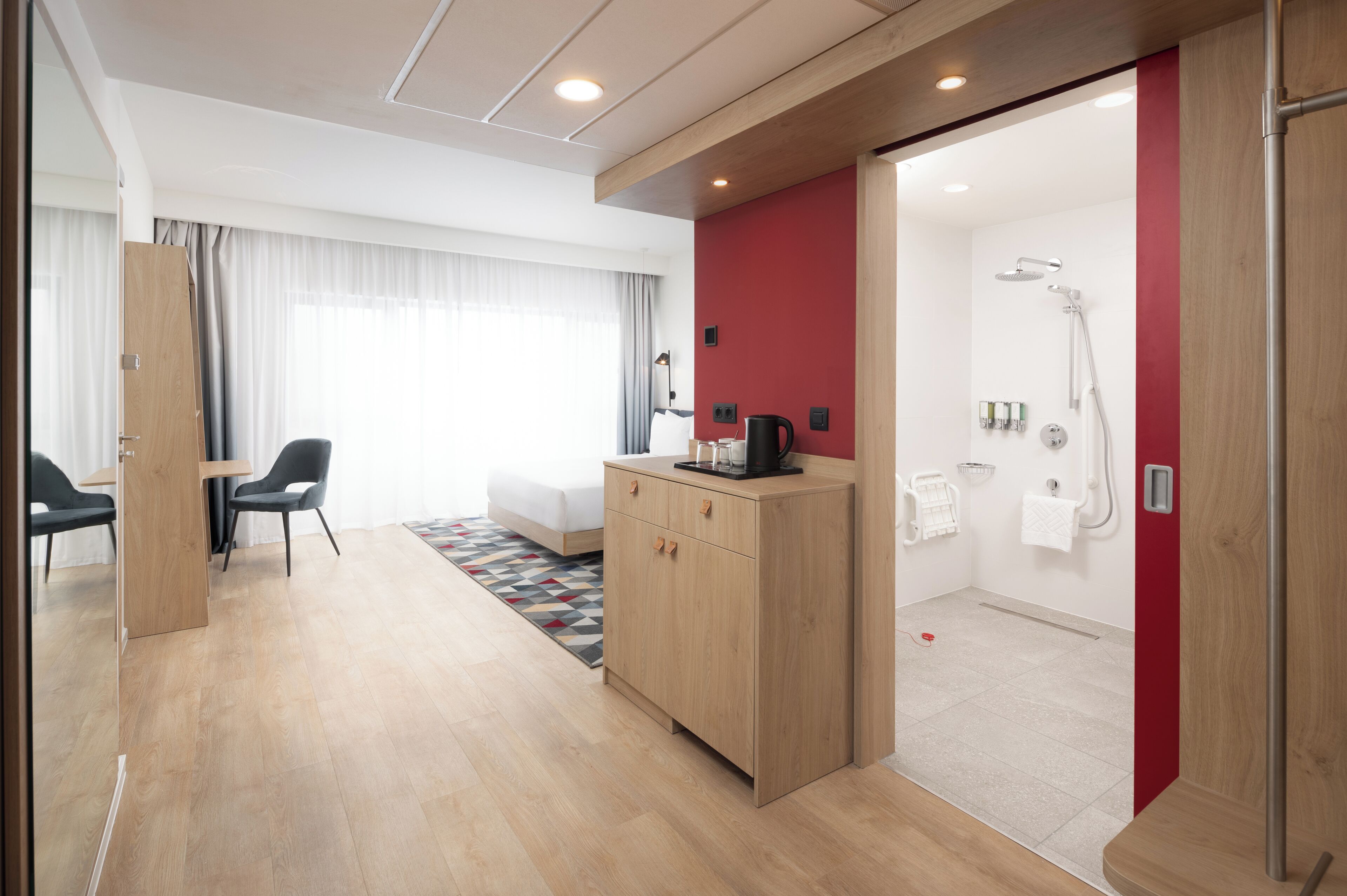 Une chambre d’hôtel moderne avec un plancher en bois, un mur d’accent rouge, un espace douche, un petit coin salon avec une table et des chaises, et un meuble en bois.