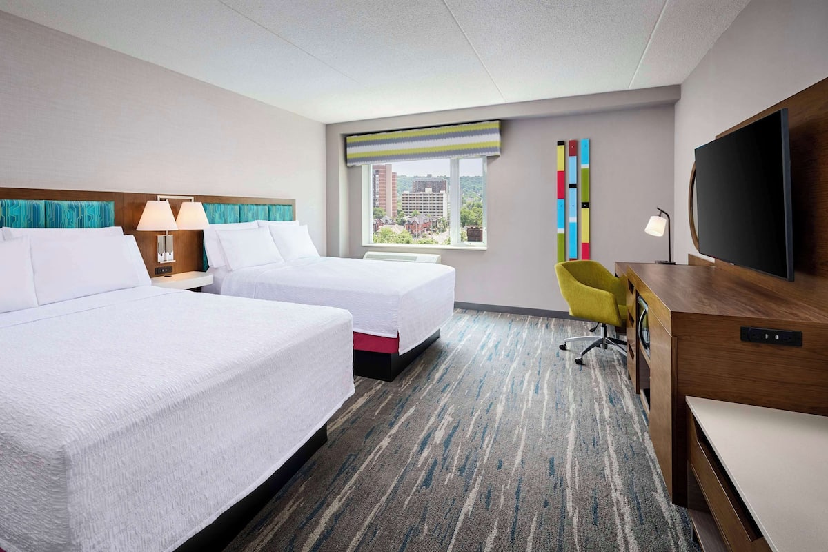 A hotel room with two beds, a desk, a television, and a window with a view of buildings.