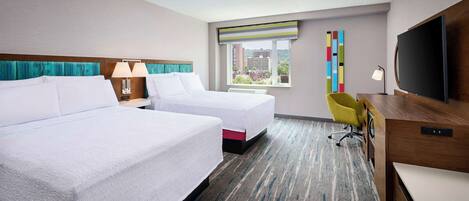 A hotel room with two beds, a desk, a television, and a window with a view of buildings.