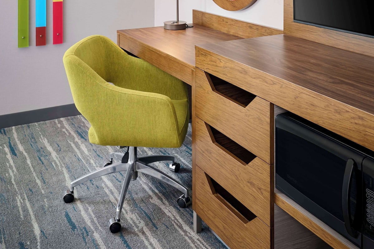 A hotel room with a wooden desk, a green chair, a flat-screen TV, a mirror, and colorful vertical blinds.