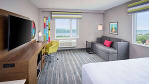 A hotel room with a large bed, a desk with a flat-screen TV, a gray sofa, and a chair. There are two windows with views of a cityscape and a large painting on the wall.