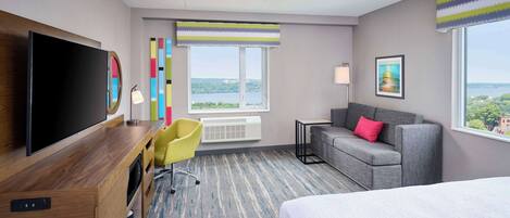 A hotel room with a large bed, a desk with a flat-screen TV, a gray sofa, and a chair. There are two windows with views of a cityscape and a large painting on the wall.