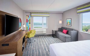 A hotel room with a large bed, a desk with a flat-screen TV, a gray sofa, and a chair. There are two windows with views of a cityscape and a large painting on the wall.