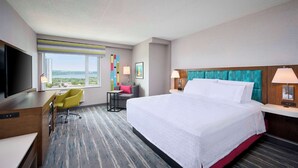 A hotel room with a large bed, a desk with a TV, a chair, a small table, and a window with a view of the city. - Hampton Inn by Hilton Hamilton (Hamilton)