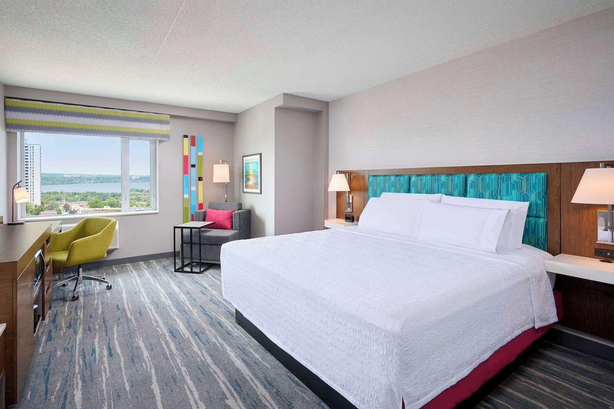 A hotel room with a large bed, a desk with a TV, a chair, a small table, and a window with a view of the city.