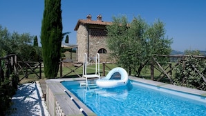 A swimming pool with a stone building in the background, surrounded by greenery and a wooden fence.