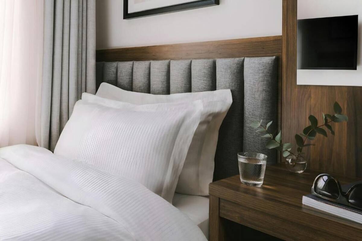 A neatly made bed with white linens, a wooden headboard, a bedside table with a glass of water, a plant, and a pair of sunglasses.