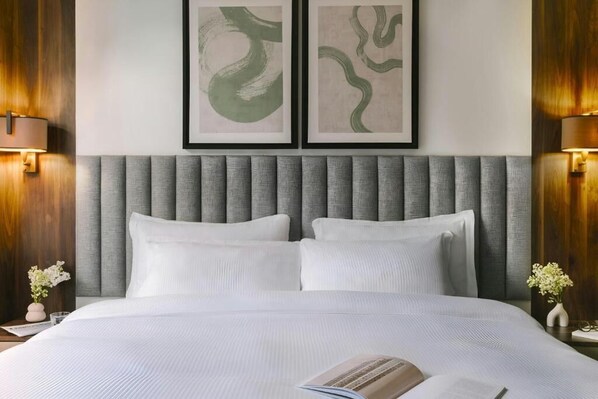 Una cama bien hecha con cabecera gris, dos cuadros abstractos enmarcados arriba y un jarrón con flores a cada lado.