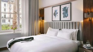 A hotel room with a large bed, a window with curtains, a nightstand with a lamp, and two framed artworks on the wall. - The Voyage Hotel Paddington  (London)