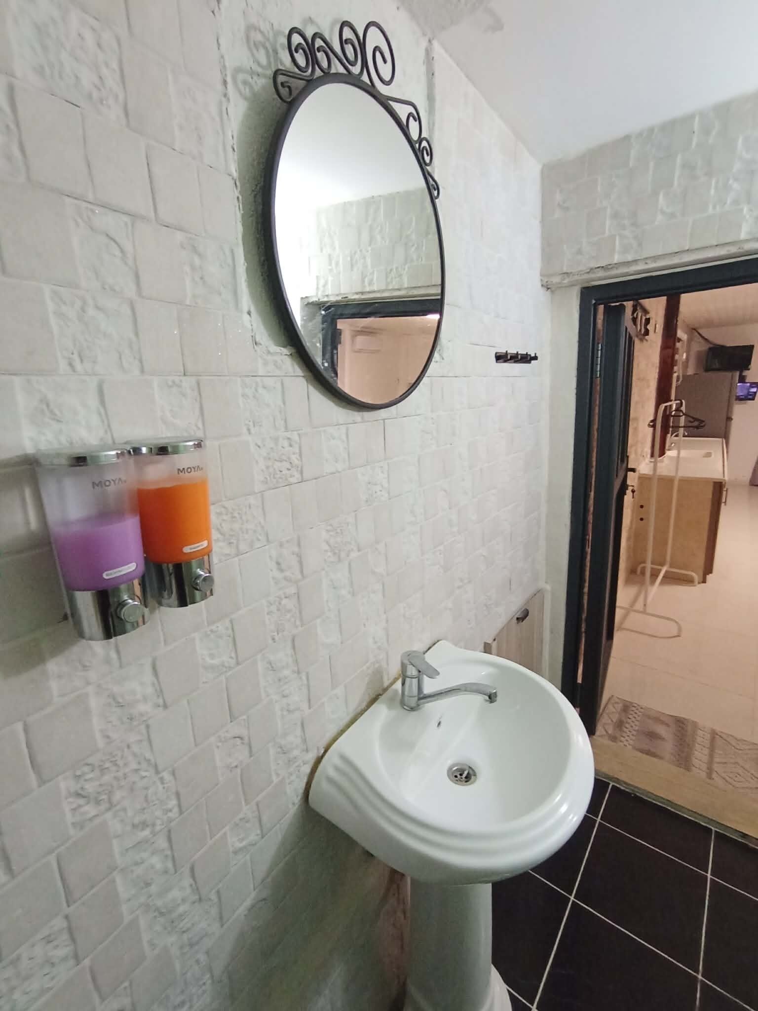 Ein Badezimmer mit weiß gefliesten Wänden, einem Standwaschbecken, einem Spiegel mit dekorativem Rahmen und einer Seifenspender.