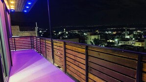 A balcony with wooden railings and a purple-lit floor, overlooking a cityscape at night.