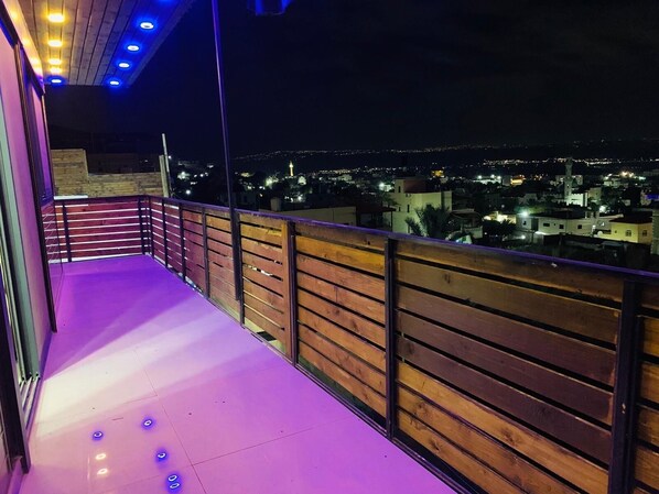 A balcony with wooden railings and a purple-lit floor, overlooking a cityscape at night.