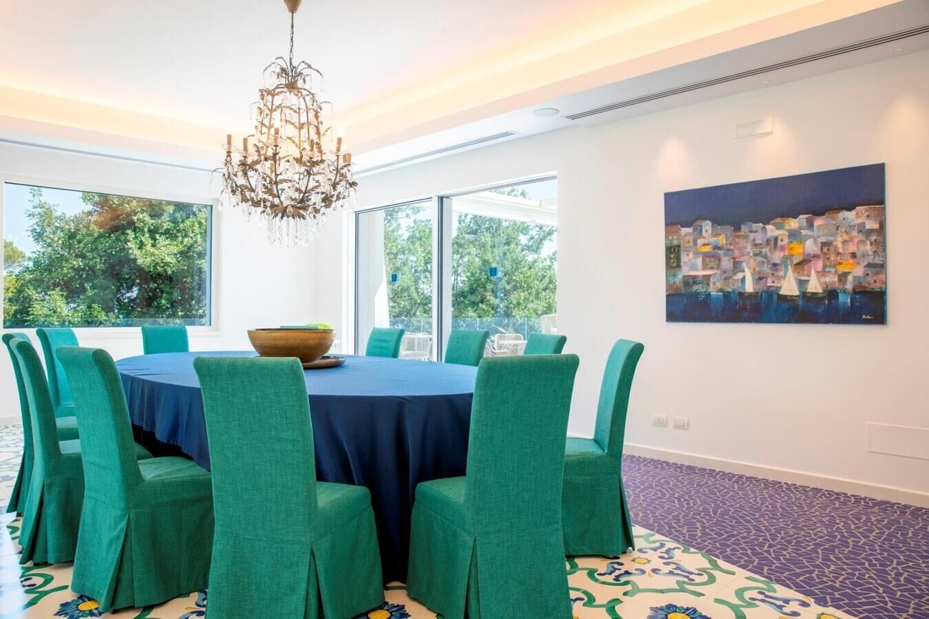 A dining area with a round table, green upholstered chairs, a chandelier, and a large window with a view of trees.
