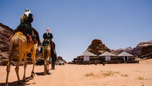 A desert scene with a camel ride, a vehicle, and a building with a unique architectural style. - Rum Crown Camp (Wadi Rum)