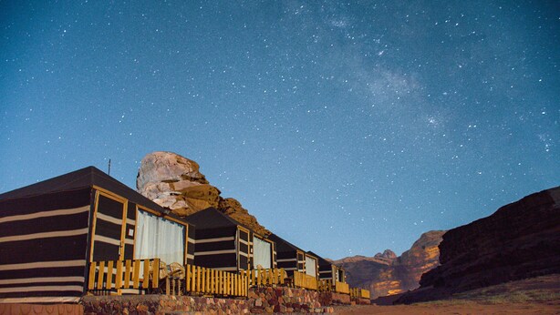 A desert landscape with a starry sky, a large rock formation, and a row of cabins with a wooden fence. - Rum Crown Camp (Wadi Rum)