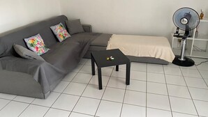 A living room with a grey sectional sofa, a small black table, a beige sofa, and a standing fan. - A 8mn de la Plage, T2 Situé au Centre de Sainte-anne Guadeloupe (SAINTE-ANNE)