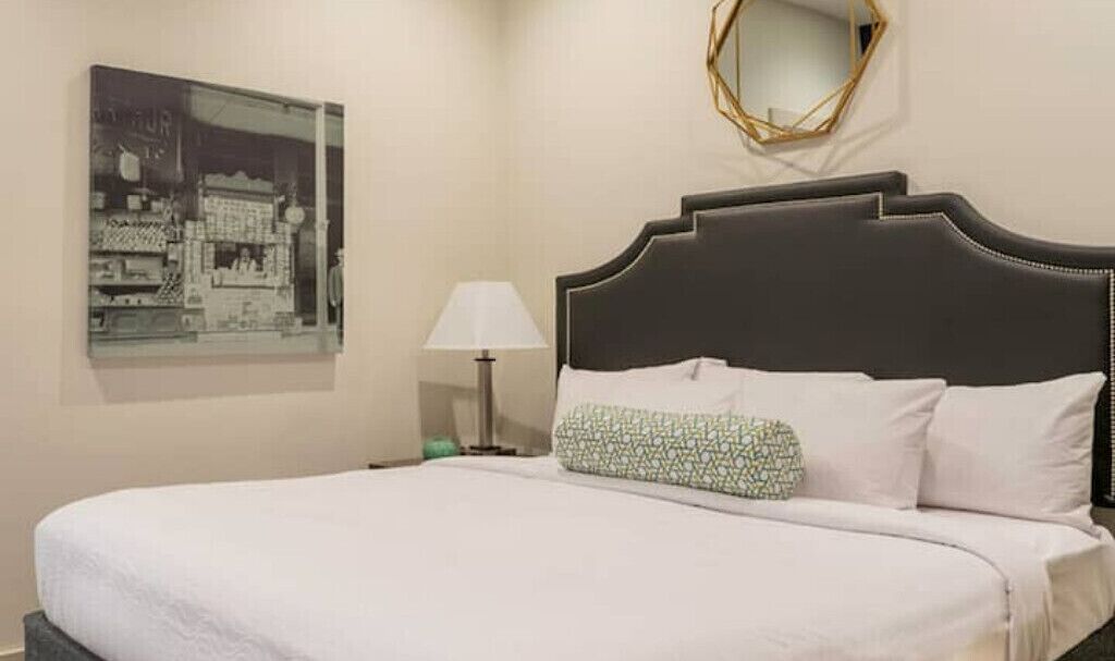 A neatly made bed with white linens, a patterned pillow, a bedside lamp, and a framed picture on the wall.