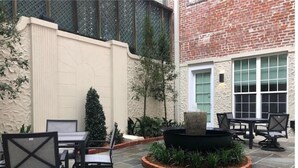 An outdoor patio area with stone flooring, black metal chairs, a round table, and a small fire pit surrounded by plants. - Unit 201 2BR Walk to Bourbon Street! (New Orleans)