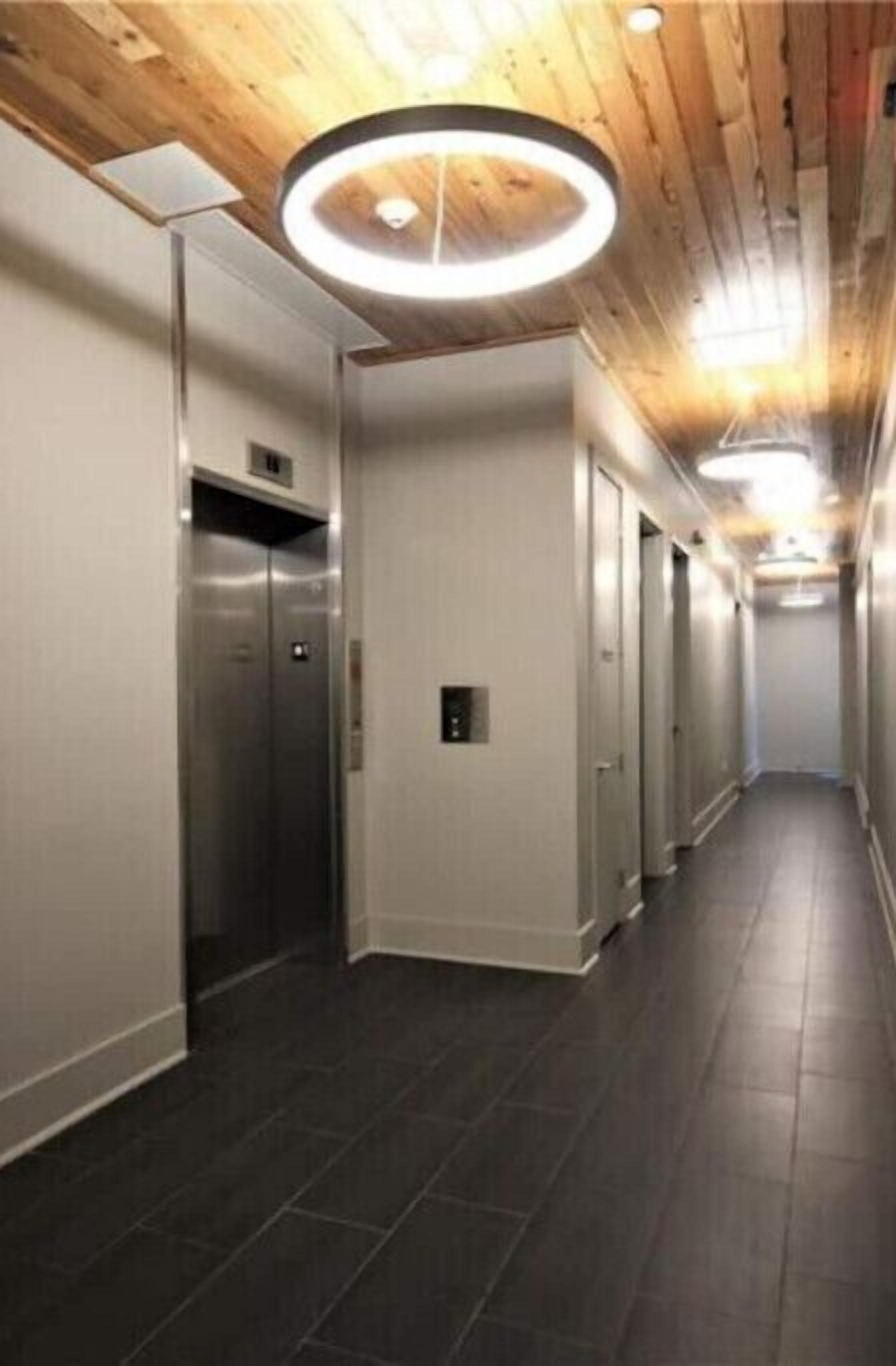 A modern hallway with a wooden ceiling, a central elevator, and a series of doors on one side.