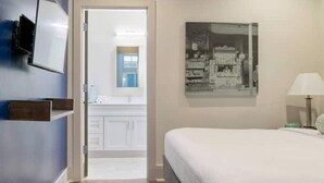 A bedroom with a bed, a nightstand, a wall-mounted TV, a painting, and a bathroom visible through an open door. - The 820 Unit 302 - 2 BR Walk to Bourbon Street! (New Orleans)