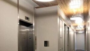 A modern hallway with a wooden ceiling, a central elevator, and a series of doors on one side.