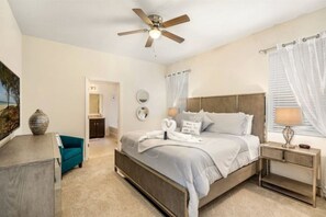 Um quarto com cama, escrivaninha, cadeira, ventilador de teto e janela com cortinas.