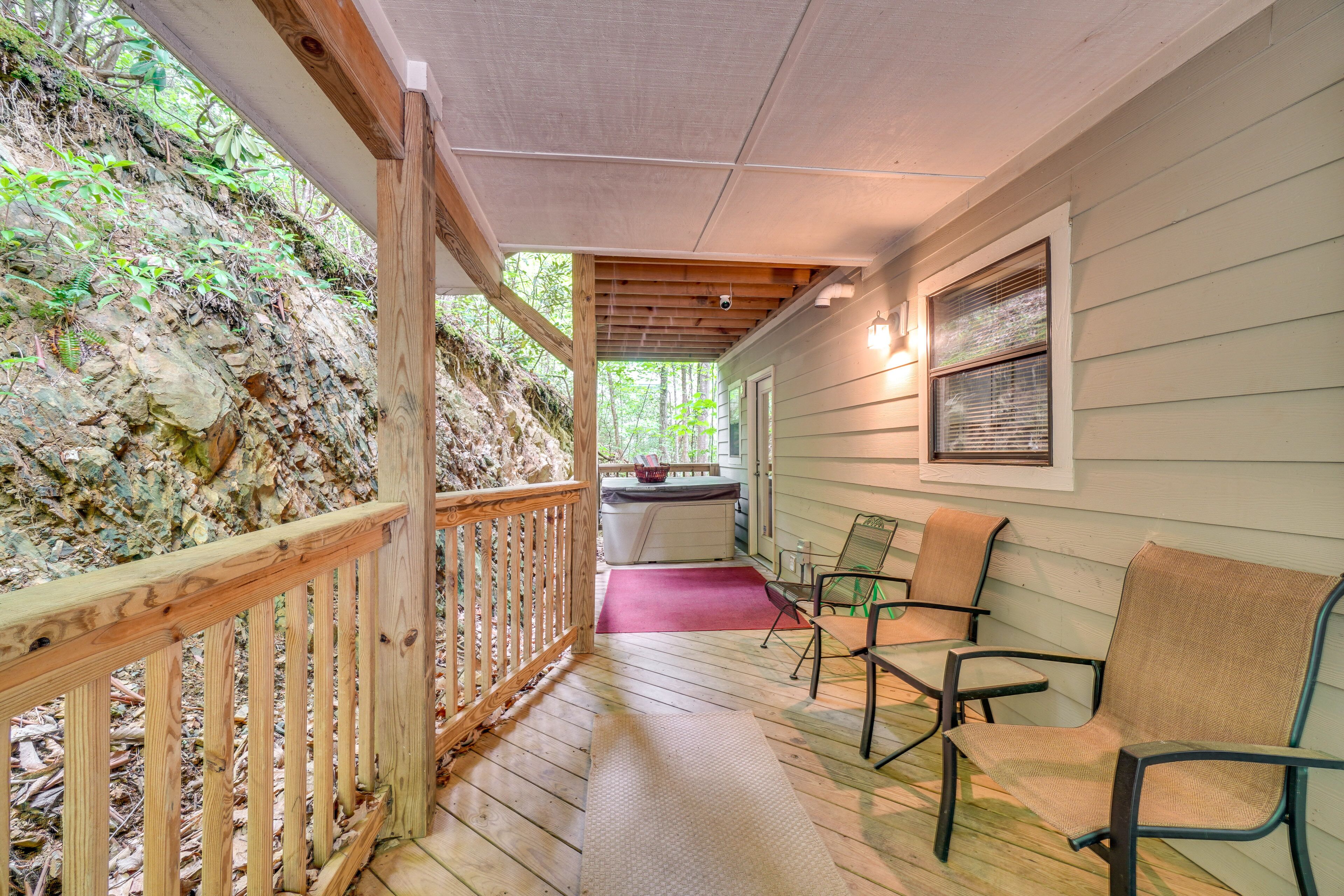 A covered porch with wooden railings, a seating area with chairs, and a view of a rocky hillside.