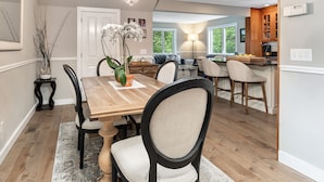 Zona pranzo con tavolo in legno, sedie e un vaso con fiori. Sullo sfondo, una cucina con mobili in legno e una finestra.