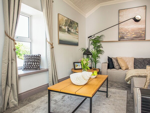 A living room with a wooden table, a sofa, a potted plant, a painting, and a window with curtains.
