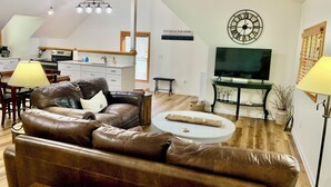 Een woonkamer met een bruine leren bank, een witte salontafel, een klok, een televisie en een keuken op de achtergrond.