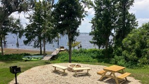 Een picknickplaats met banken en een vuurkuil, omringd door grind, met uitzicht op een waterpartij met boten en bomen.