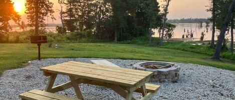 A picnic table and a fire pit in a park with a lake and trees in the background.