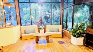 Une pièce chaleureuse avec un plancher en bois, un canapé beige agrémenté de coussins à motifs, une table basse en bois et une grande fenêtre donnant sur des arbres et un immeuble.