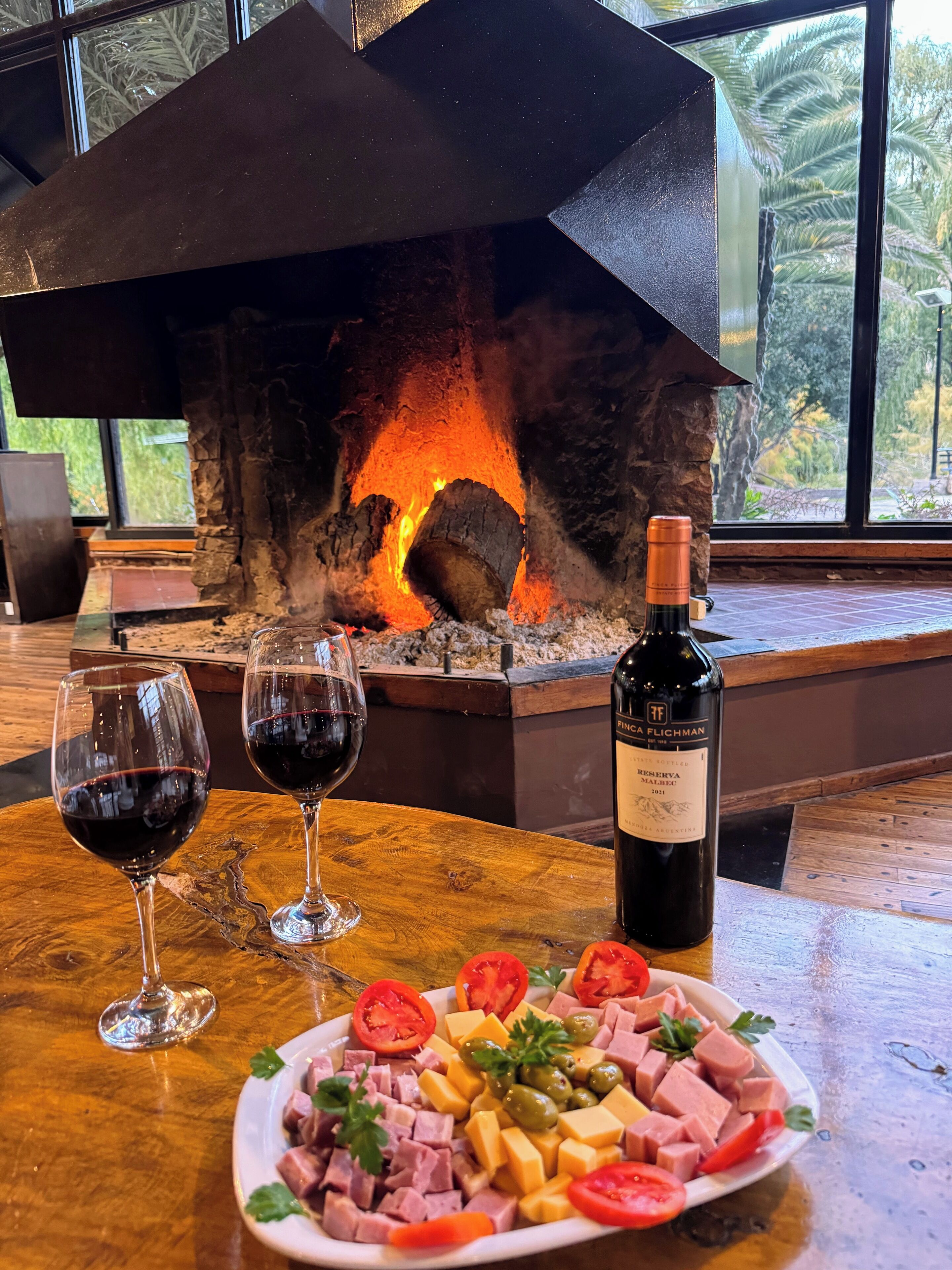 A fireplace with a fire, two glasses of red wine, a bottle of wine, and a plate of food.