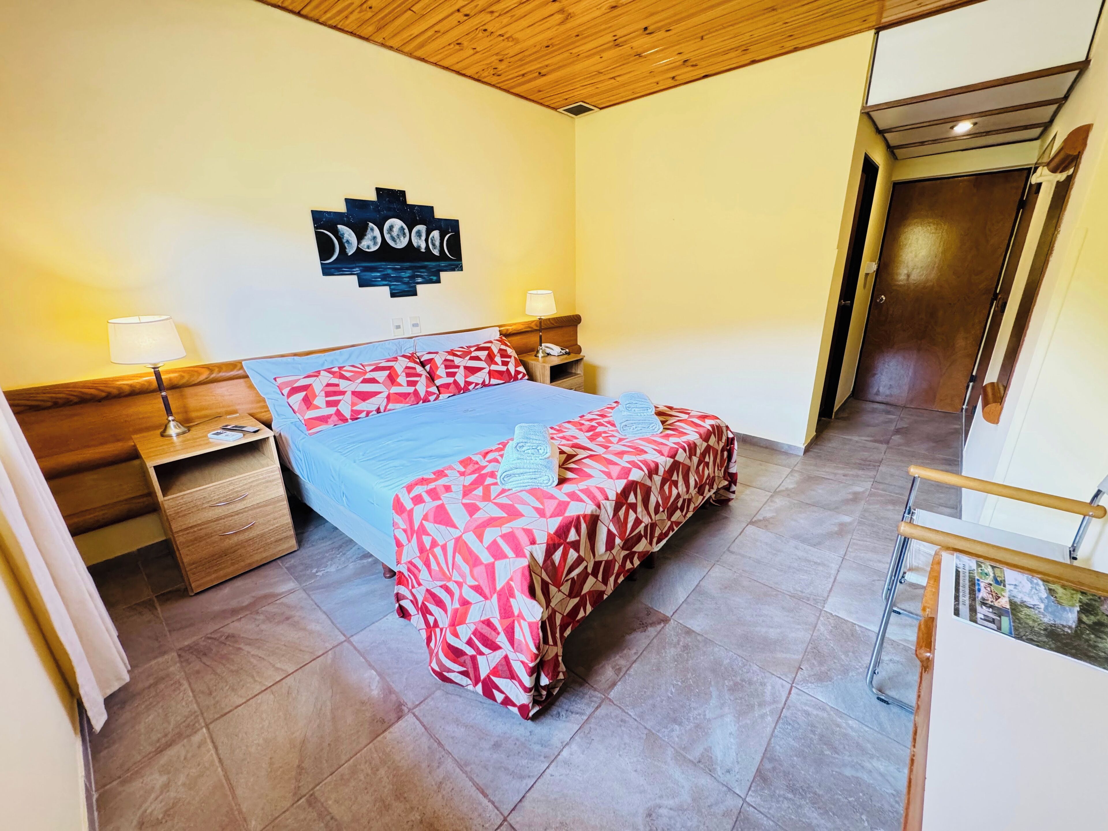 A bedroom with a bed, bedside tables, a nightstand, a wooden headboard, a wall art piece, and a door.
