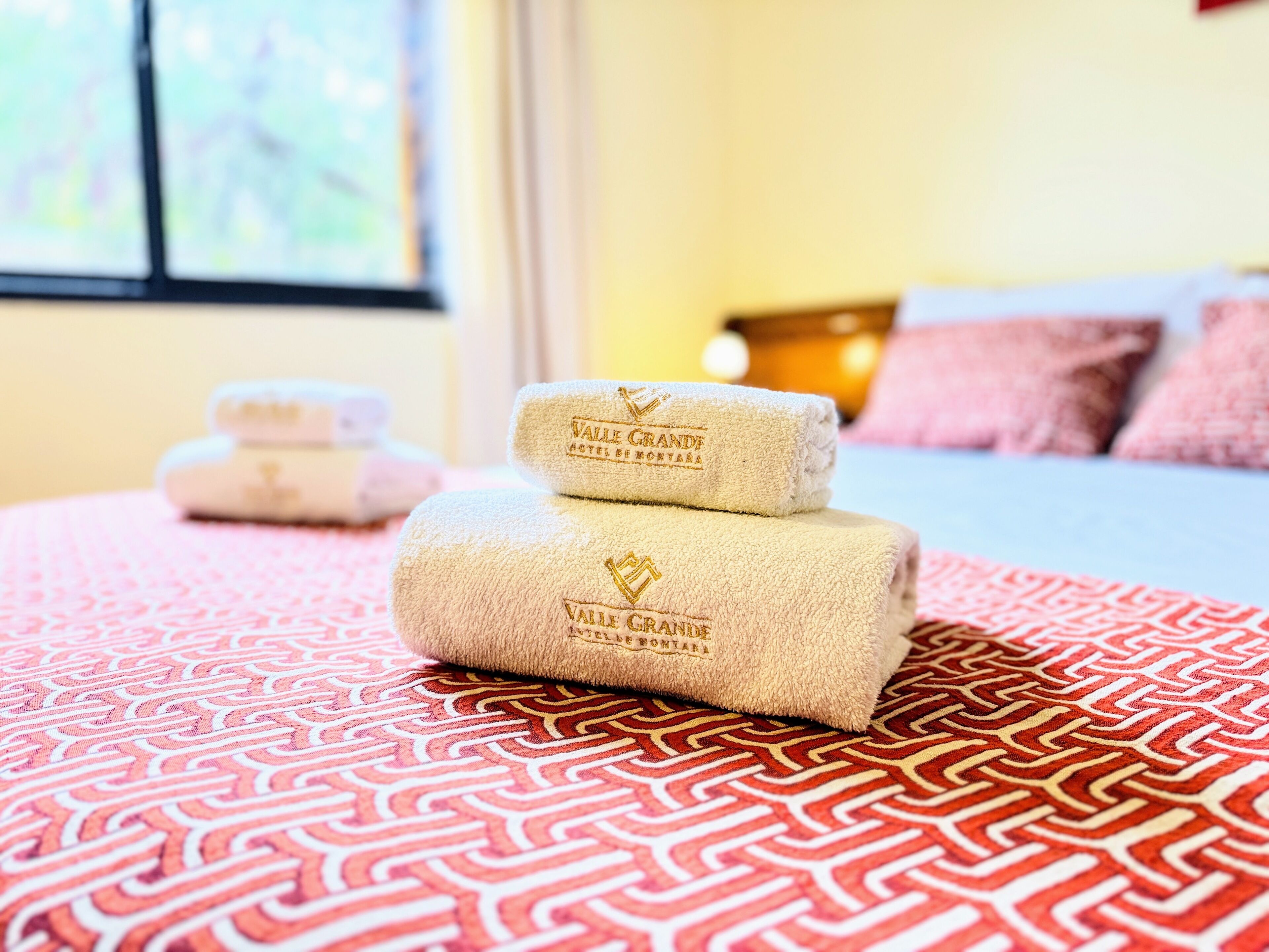 A neatly made bed with a patterned bedspread, two rolled towels with the hotel name, and a window with a view in the background.