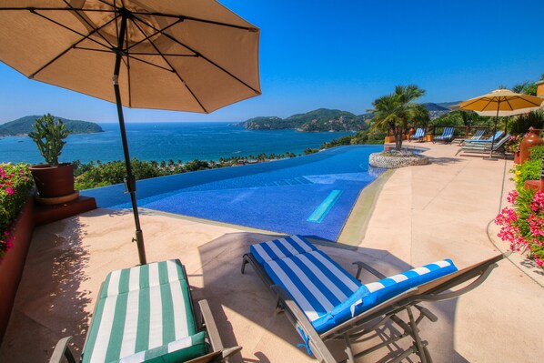 Zona de piscina con sillas de descanso y sombrillas, con vista al mar azul turquesa y colinas lejanas.