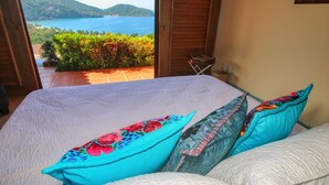 Una cama con colcha blanca y almohadas coloridas, una puerta de madera y vistas a un cuerpo de agua y vegetación.