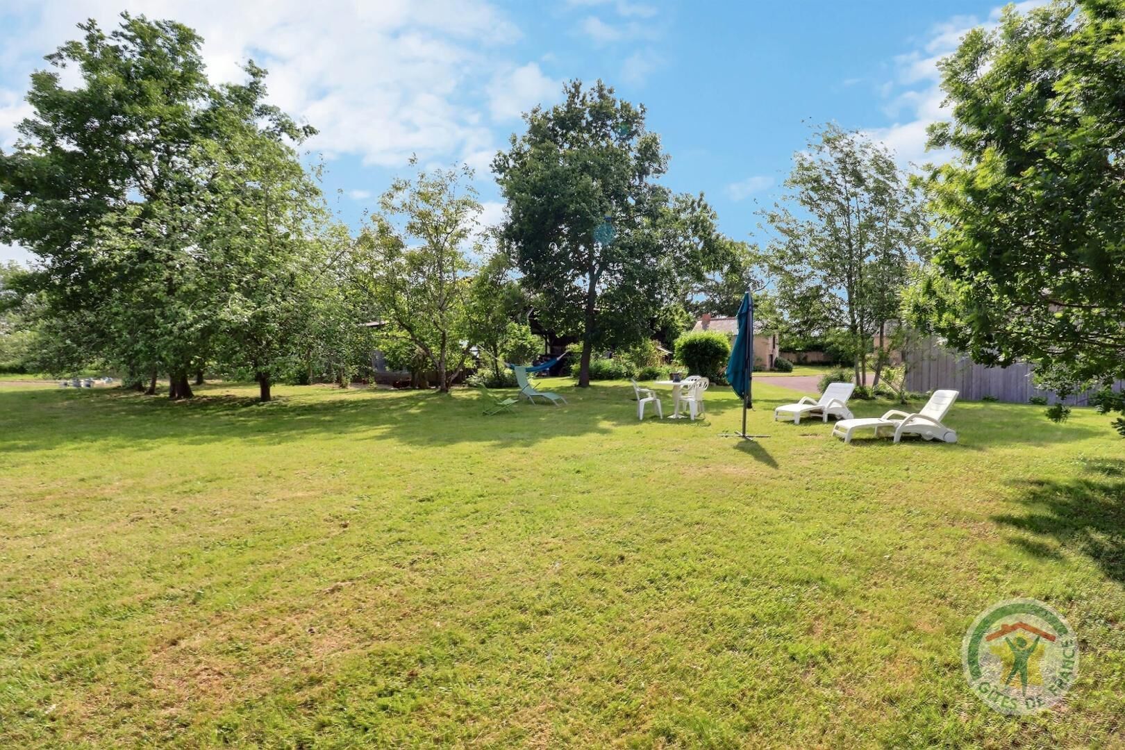 Un espacio al aire libre con césped, sillas reclinables blancas y una mesa, rodeado de árboles.
