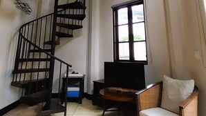 Ein großzügiger Wohnbereich mit Wendeltreppe, einem Flachbildfernseher, einem Holzstuhl und großen Fenstern, die für natürliches Licht sorgen.